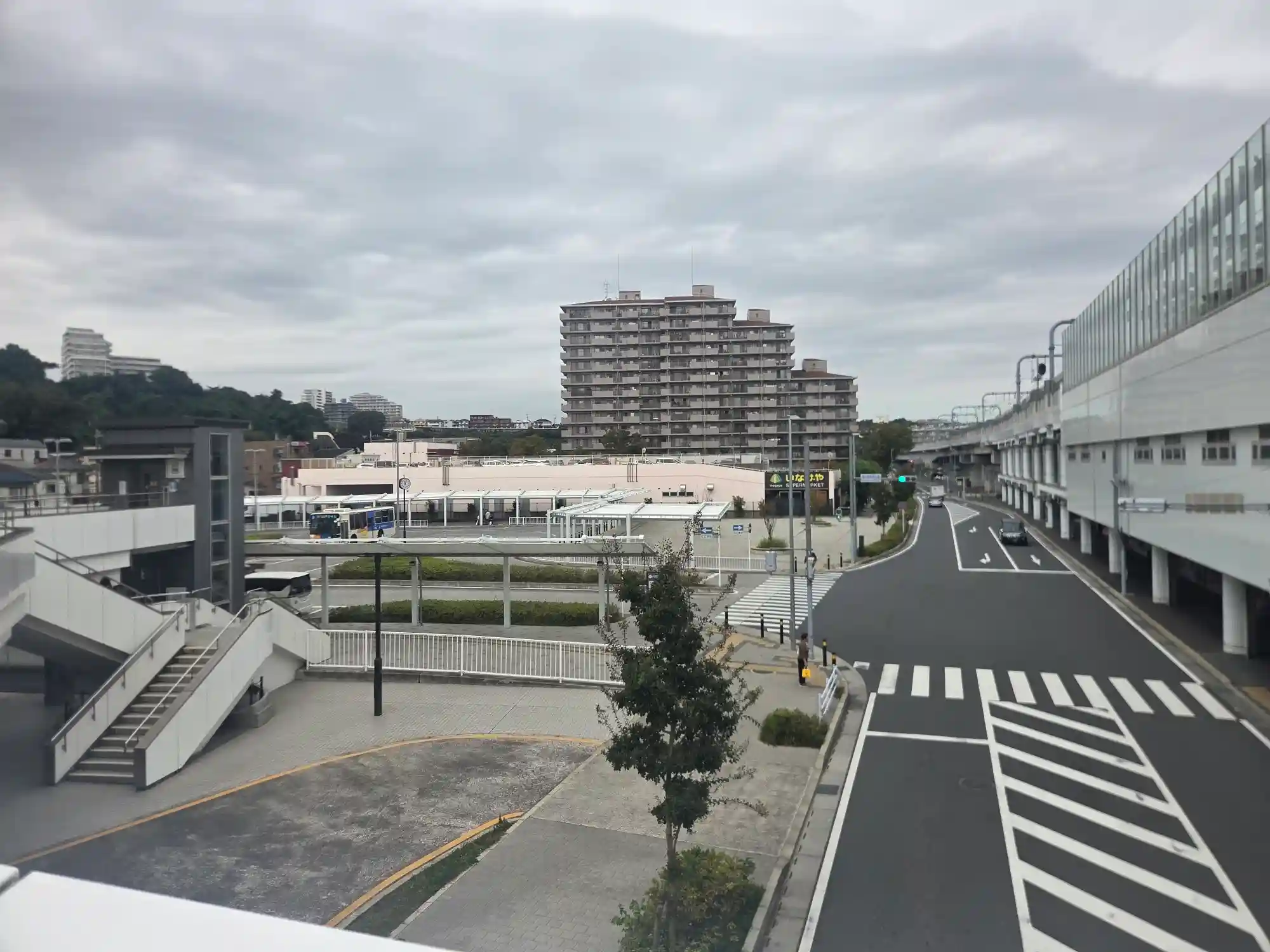 星川駅前のバスロータリーを歩道橋から見た様子。奥にいなげやと大型マンション、左奥に緑の多い丘が見える