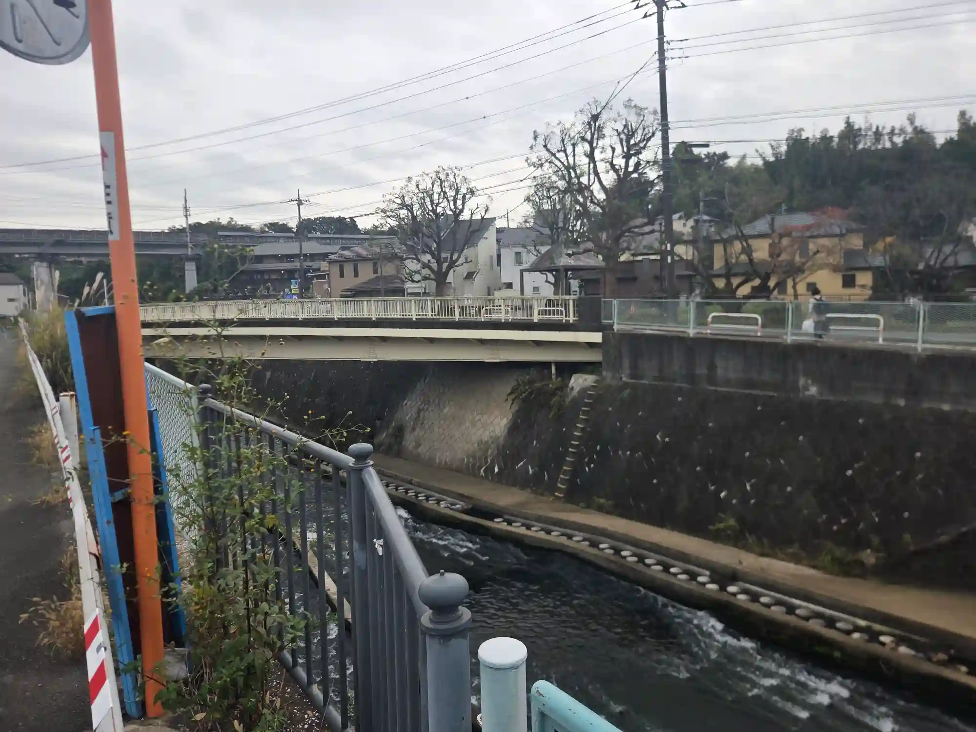 西谷駅近くの川と橋。川沿いの低地の奥に、木に囲まれた丘の上の住宅が見える