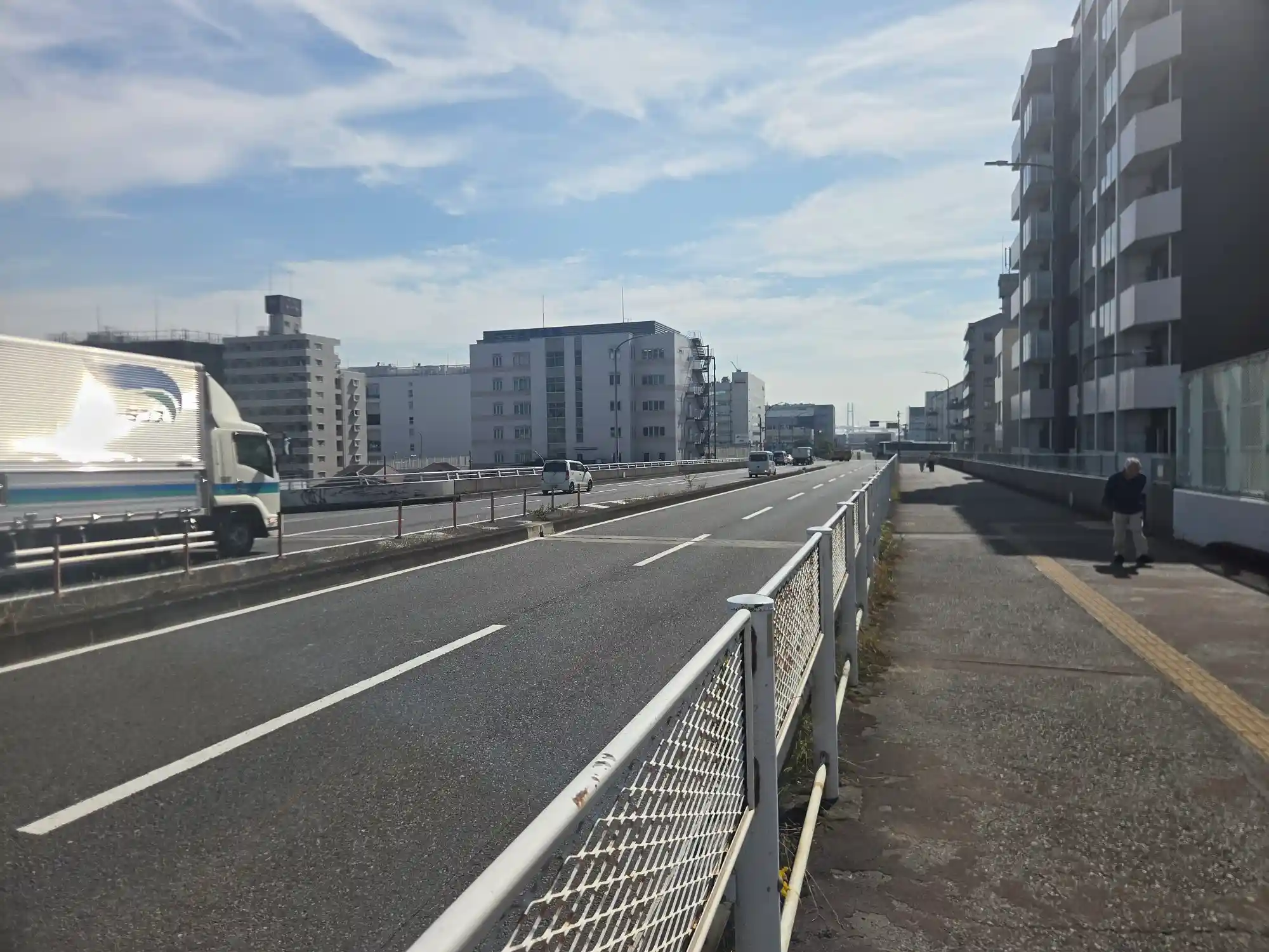 新子安駅南側の幹線道路と歩道。大型トラックが走り、沿道には新しいマンションとオフィスビルが並ぶ