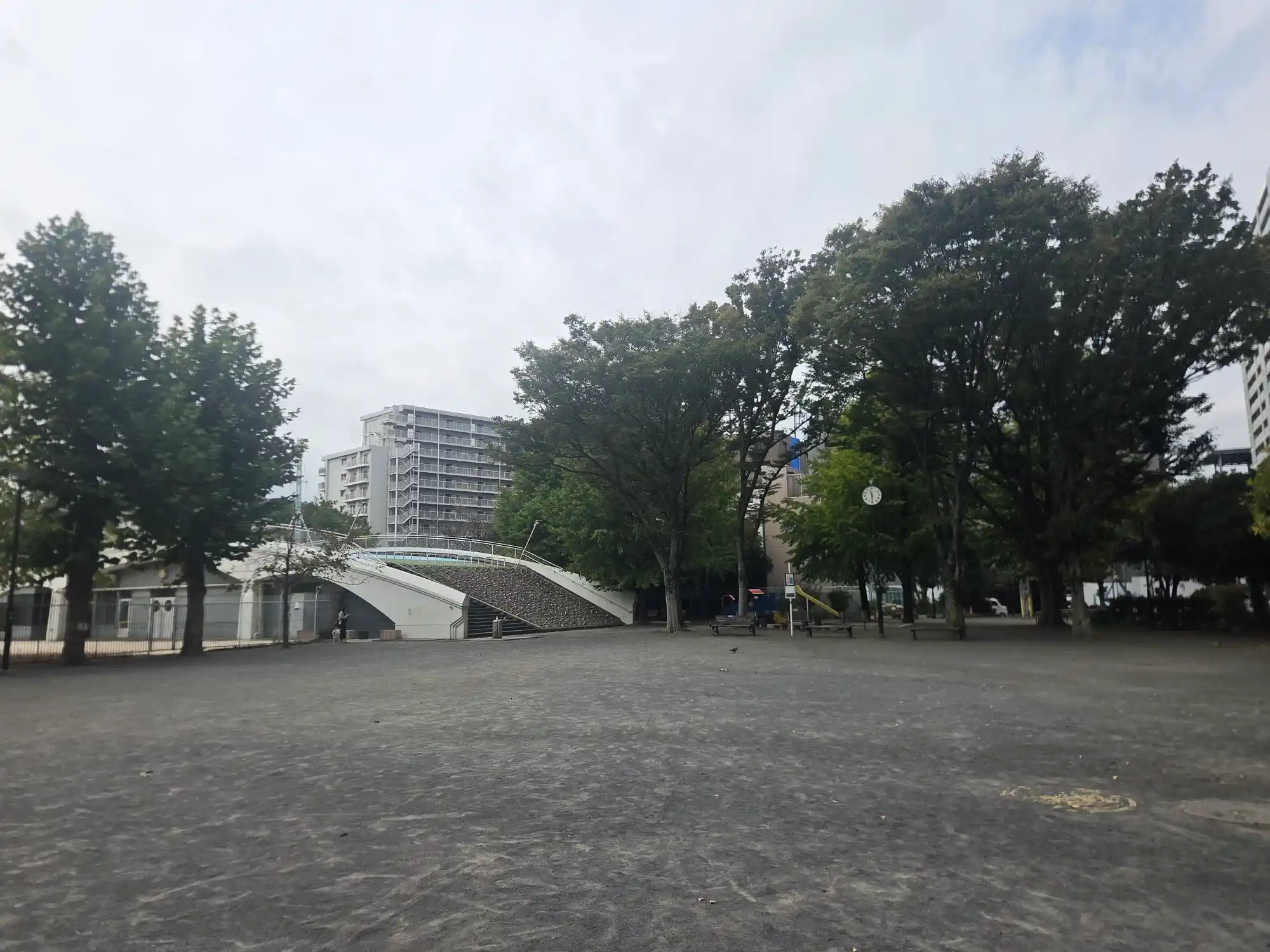 天王町駅近くの平坦な広い公園。大きな木の下にベンチと時計があり、奥にマンションとアーチ形の階段がある